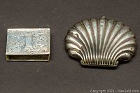 Top view of sterling silver matchbox case next to shell-shaped silver powder compact on black background.