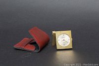 Sweda travel clock and its red leather pouch displayed separately on a black surface showing the clock face and metal casing.