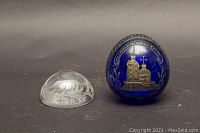 Two paperweights on a dark background. One is a clear etched glass dome marked Edinburgh Scotland. The other is an egg-shaped cobalt blue paperweight with gold etched Russian church imagery and Cyrillic script.