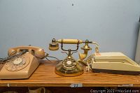 Three vintage telephones on wooden shelf against blue wall: beige Northern Telecom rotary phone, brass French style rotary phone, and beige Uniden cordless phone