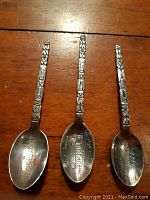 Three vintage sterling silver spoons showing detailed totem pole motifs on handles and engraved bowl interiors, laid flat on wooden surface.
