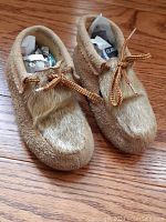 Pair of light brown suede moccasins with fur tops and yellow lace ties on wood floor.
