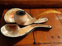 Photo of vintage meerschaum pipe resting in original fitted case on wood surface showing pipe bowl and amber curved stem.