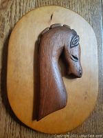 Front view of hand carved teak war horse relief on oval wooden plaque showing horse head profile and wood grain texture.