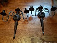 Two bronze powder coated iron candelabra wall sconces placed side by side on wooden floor showing overall design and size.