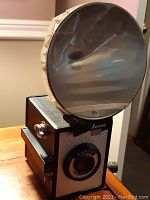 Side angled view of the Ansco Shur-Flash box camera with attached large round flash reflector, showing camera body and flash size.