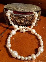 Photo showing two pearl bracelets draped over an ornate Victorian style metal trinket box with red interior.