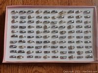 Box of 100 assorted silver-tone fashion rings arranged in rows on a white insert inside a flat pink box.