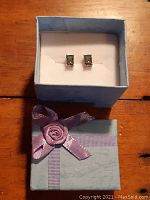 Pair of emerald cut ocean blue Mystic Topaz stud earrings, set in sterling silver, displayed in a light blue gift box with a decorative pink ribbon and flower on the lid.
