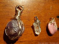 Three stone and crystal pendants laid on wooden surface; includes purple amethyst wire wrapped, clear quartz with gold claw setting, and pink stone with gold claw setting.