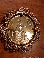 Close-up photo of 1964 gold plated British one penny coin in ornate filigree holder, showing reverse with seated Britannia detail.