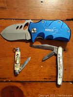 Three vintage pocket knives displayed on wood surface: blue Boker Plus folding knife with wide blade, small cream handled knife with woman image and one blade, white handled Mullite Tint & Awning Co. knife with two blades.