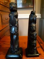 Photo showing two black stone totem poles with carved eagle and beaver figures, standing upright on a wooden surface.