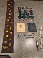 Brown sash laid flat showing 14 embroidered yellow and brown badges along with 8 round badges placed beside it, several pins, a booklet, silver brooch, whistle with cord, and two safety pins.