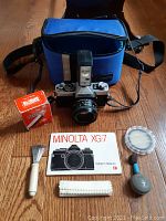 Full view of Minolta XG7 camera with lens and flash, blue carrying case, boxed flash bulb, manual, and cleaning tools arranged on wooden floor.