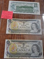 Two Canadian one dollar bills from 1973, last issue, placed inside protective plastic sleeves. Bills have sequential serial numbers BFG4136258 and BFG4136259. Both show the front with the Queen Elizabeth II portrait and the Bank of Canada text.