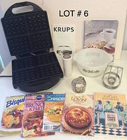 Photo shows KRUPS waffle maker open with square waffle grids, six assorted cookbooks standing upright, two metal biscuit cutters, a white glass mixing bowl with a green floral pattern, and a white powder sugar shaker arranged on surface.