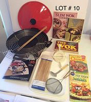 West Bend electric wok with red lid, metal rack inside, chopsticks laying on the rack, skewers in packaging, a plastic dumpling press, a skimmer spoon, a ladle, and five assorted cookbooks arranged on a white surface.