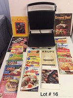 Photo of the Krups Universal Grill & Panini Maker and 17 related cookbooks arranged on a surface with titles visible related to grilling, hamburgers, and ground beef recipes.