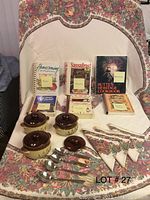 Photo showing assorted state cookbooks, three ceramic bowls with lids, six metal spoons, a floral patterned tablecloth and matching napkins.