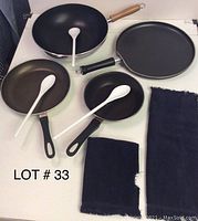 Four Teflon cooking skillets with black and wooden handles, three white plastic spoons, and two dark cloths on a surface.