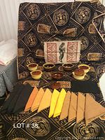 7 ceramic bowls in brown and yellow tones, assorted colorful cloth napkins, and a patterned tablecloth with tan and black geometric designs.
