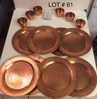 Seven copper chargers arranged in two rows on a table, with five copper candle holders and a white plastic box with small candles at the top