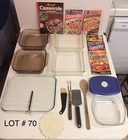 Photo shows five Pyrex glass dishes in amber and clear glass, one clear Pyrex glass square platter with grid markings and a thermometer on it, four kitchen utensils including a basting brush, two forks, a metal grater, and a wooden spoon, and six casserole cookbooks with colorful covers arranged behind the dishes.