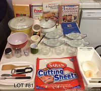 Photo shows assortment of kitchenware including glass and ceramic bowls, metal serving dish, five cookbooks, and various utensils and tools.