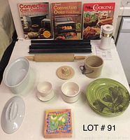 Photo of lot items showing ceramic baking dish, bread loaf pan with lid, egg cups, crock jar, rolling pin, butter dish, green plate with floral design, and three cookbooks.