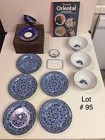 Five blue floral patterned plates, three white bowls with blue fish motif, small dish, dragon cup and saucer on wooden crate, and cookbook.
