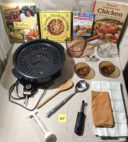Electric grill, copper Corning Ware amber glass saucepan, two amber bowls, cookbooks, utensils, towels arranged on table.