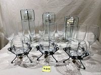Photo showing 3 clear glass bowls on black wire holders with grape decoration in front, 3 rectangular glass candle holders with black metal frames in back, and 9 clear wine glasses arranged between them.