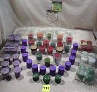 Lot of assorted color votive candles arranged on a flat surface with neutral background. Colors include green, orange, red, purple, and white. Some candles are in glass holders and some are boxed. There are also several empty clear glass holders behind the candles.