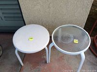 Photo shows two small round stackable outdoor tables side by side. One table has a solid white top, and the other has a clear textured glass top with white metal legs on a concrete surface.