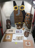 Collection of owl-themed items displayed including a woven owl rug, wooden wall holders, framed and unframed owl art prints, a small basket with soft owl sculptures, and wooden owl cut outs