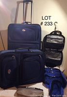 Photo showing two rolling suitcases, one large suitcase with extendable handle and missing zipper slider in front pocket, medium suitcase, hanging toiletries bag, and four blue storage bags on floor.