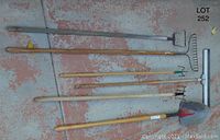 Five assorted garden tools laid out on ground, including two rakes, a hand cultivator, a shovel, and a long handle water squeegee