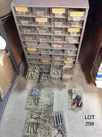 Grey parts bin with multiple small drawers, some missing, each containing various hardware such as bolts, nuts, and washers spread out in open drawers and on the floor in front of the bin.