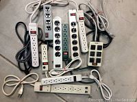 Top-down photo showing assortment of pre-owned power strips in white and black colors with varying outlet counts and lengths, with cords tangled together.