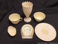 Six Belleek porcelain pieces displayed on black fabric: goblet, cup, saucer, two bowls, ashtray shaped like horseshoe with shamrocks.