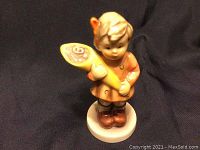 Front angle of the Hummel figurine showing child holding yellow cone on round base against dark background.