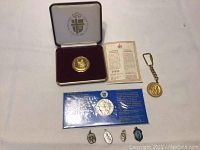 Lot contents including medals, keychain, pendants, certificate, and packaging arranged on white fabric background.