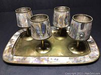Four brass and abalone shell goblets placed upright on the matching brass tray with abalone shell border, showing iridescent shell patterns and brass surfaces.