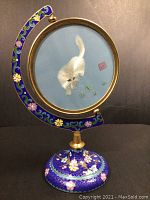 Front view showing white cat on blue silk with grasshopper detail, mounted on dark blue floral cloisonne swivel stand with brass rim.