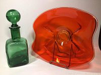 Photo showing green square glass decanter with round ball stopper and orange wavy blown glass bowl on display stand.
