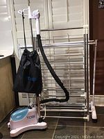 TOBI upright portable steamer with travel bag, hose attached, positioned next to drying racks on casters.