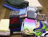 Multiple stacks of assorted towels, washcloths, blankets, and comforters including Disney printed towels and various patterned and solid colored linens on floor