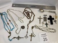Photo showing multiple rosaries made of different materials, metal crosses, religious medals, and a silver spoon marked Kowloon, all displayed with a ruler for scale.