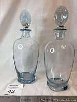 Pair of light blue hand blown glass decanters with stoppers, standing side by side against a white backdrop.